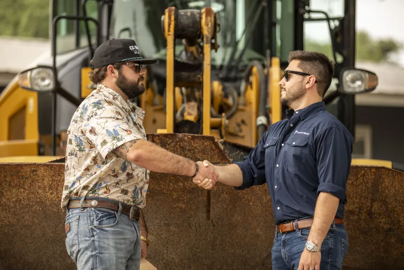 Handshake in front of a raised loader bucket — deal done