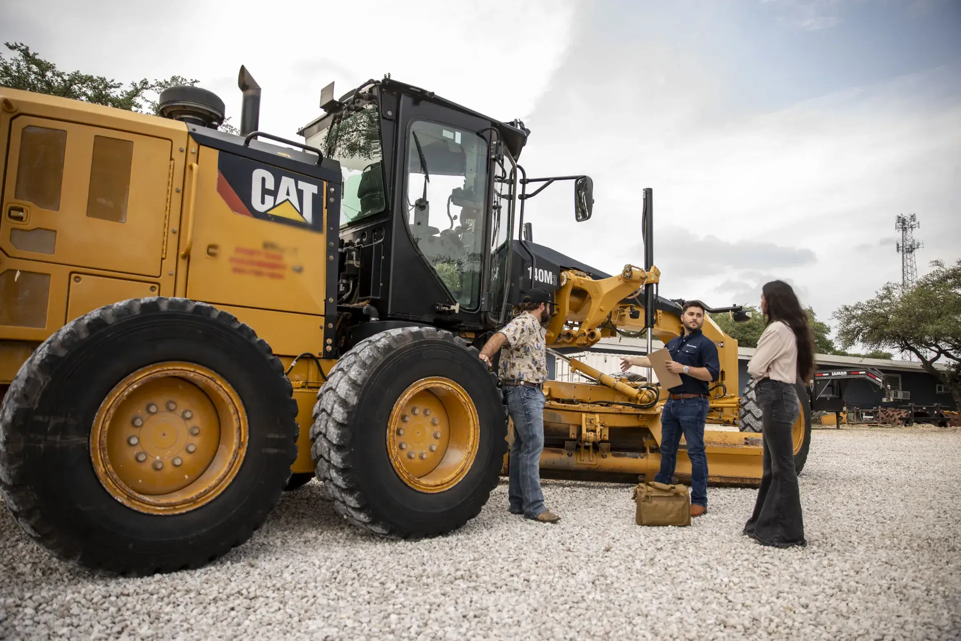 Three people walking a motor grader on a gravel lot, catalog in hand